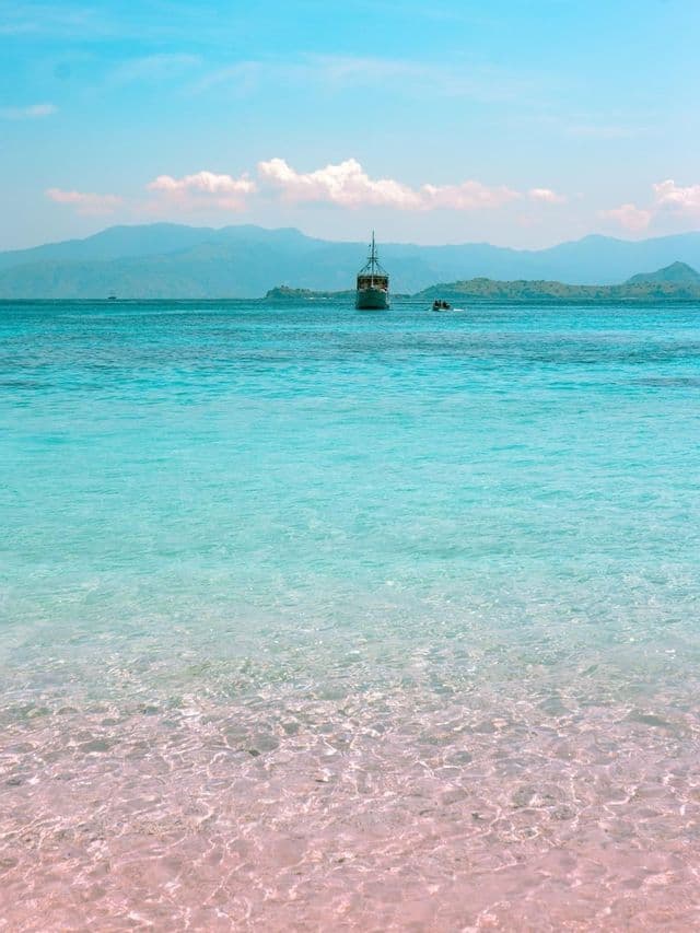 Una barca di legno ancorata in acque limpide e turchesi al largo di una spiaggia di sabbia rosa, con montagne sullo sfondo sotto un cielo blu.