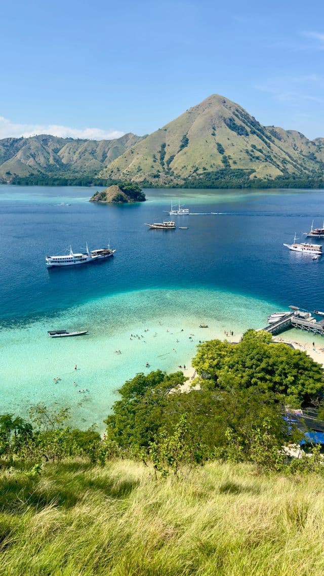 Una vista aerea di una baia tropicale con persone che nuotano in acqua turchese, barche a vela e isole verdi sullo sfondo.
