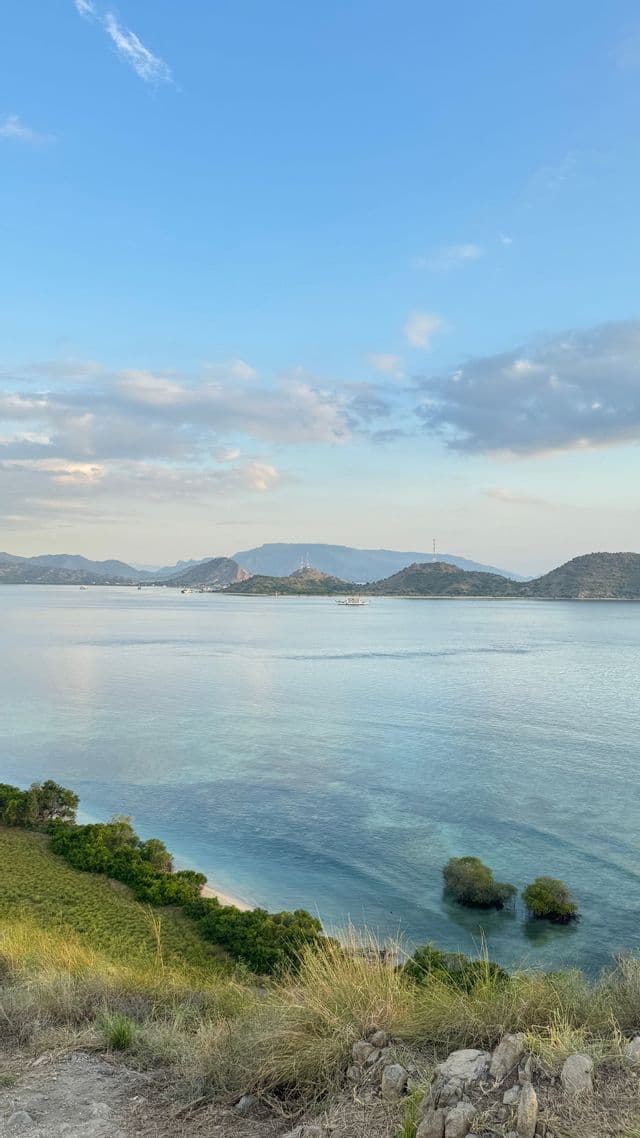Una vista da una collina erbosa che domina una baia calma e turchese con isole lontane sotto un cielo blu con nuvole sparse.