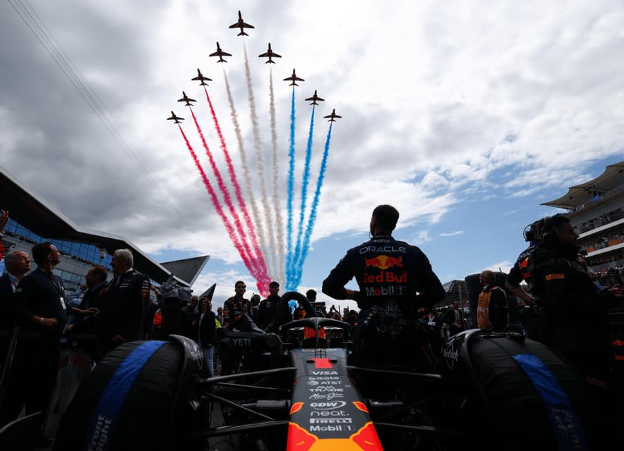 From behind a Formula 1 car, a driver and crowd watch jets fly in formation, leaving red, white, and blue smoke trails in the sky.