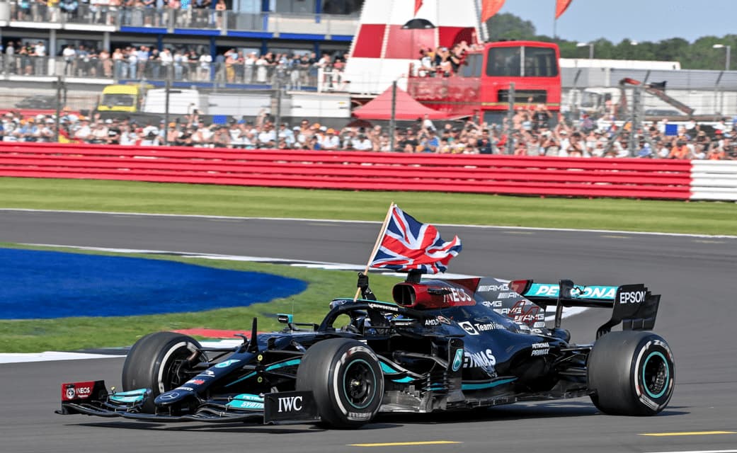 A Formula 1 driver in a black race car holds up a British flag while driving on a racetrack in front of spectators.