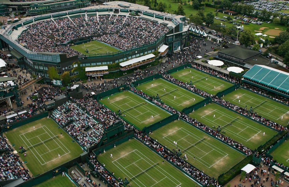 Aerial view of a packed tennis stadium and numerous grass courts with ongoing matches and crowds of spectators.