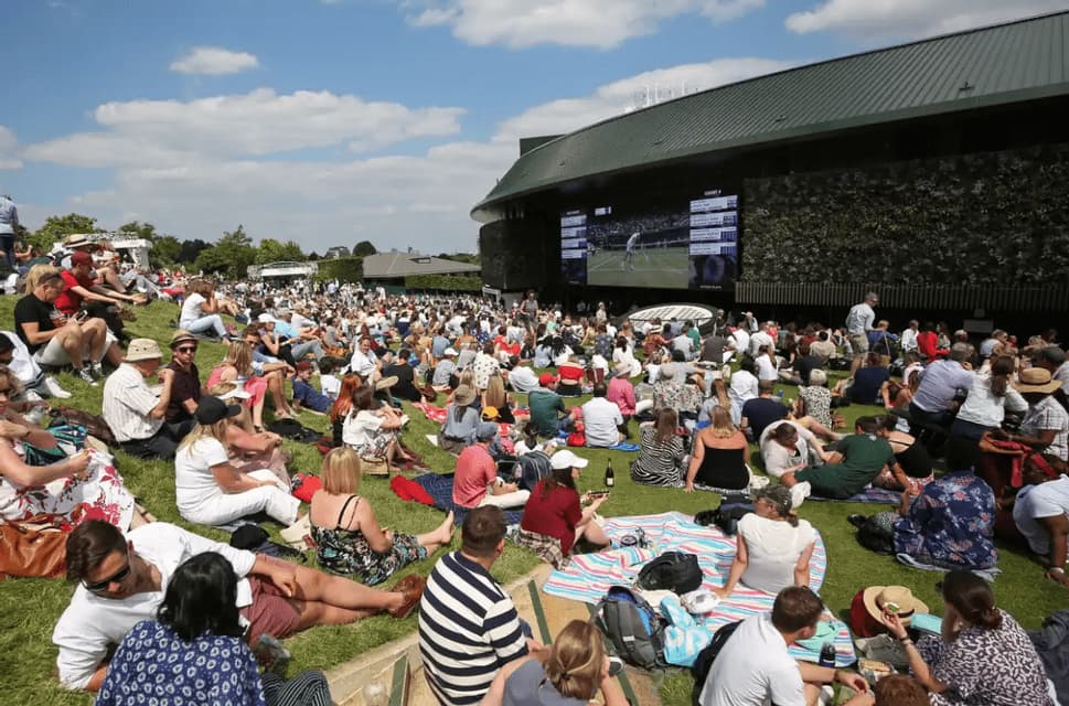 A large crowd of people sits on a grassy hill watching a tennis match on a giant outdoor screen on a sunny day.