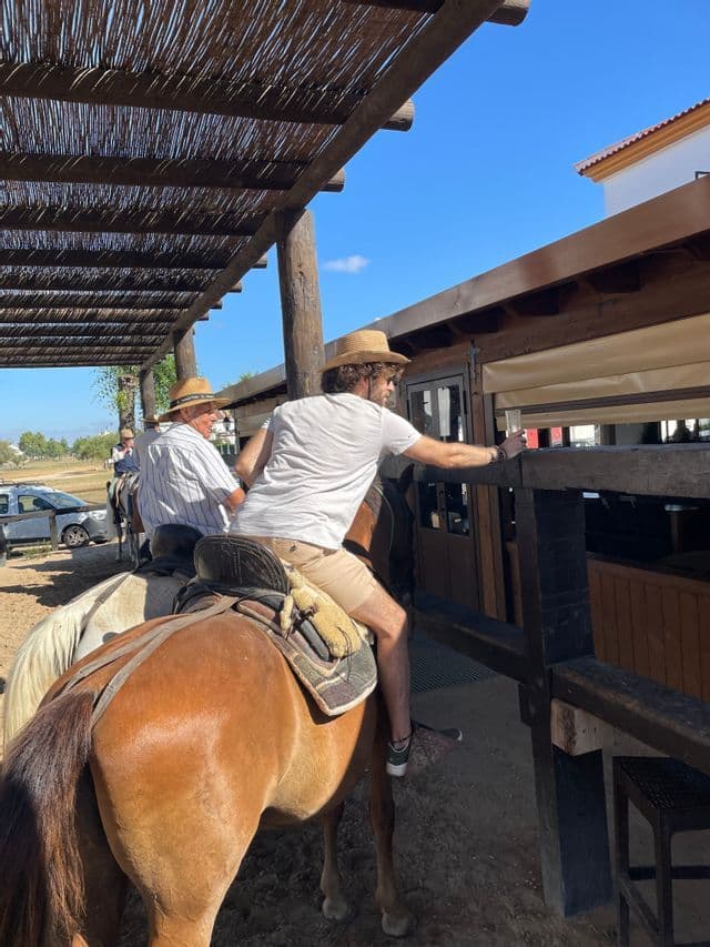 Un uomo di un viaggio di gruppo WeRoad a cavallo si protende per un drink in un bar di legno all'aperto sotto una pergola.