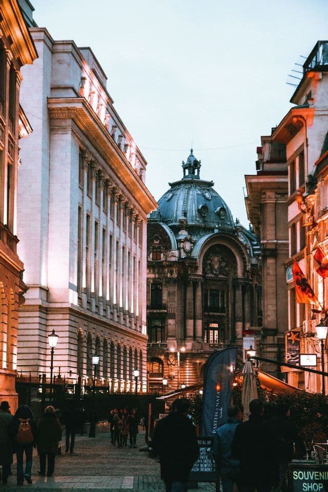 An ornate building with a large dome stands at the end of a cobblestone street where people are walking at dusk.
