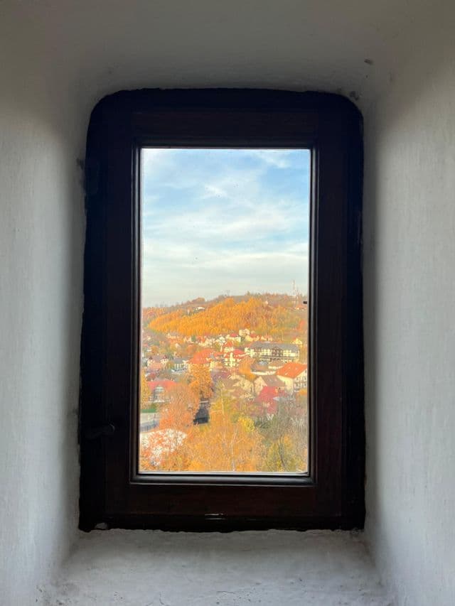 Veduta di un borgo collinare con alberi autunnali arancioni, vista attraverso una finestra di legno scuro da un interno.