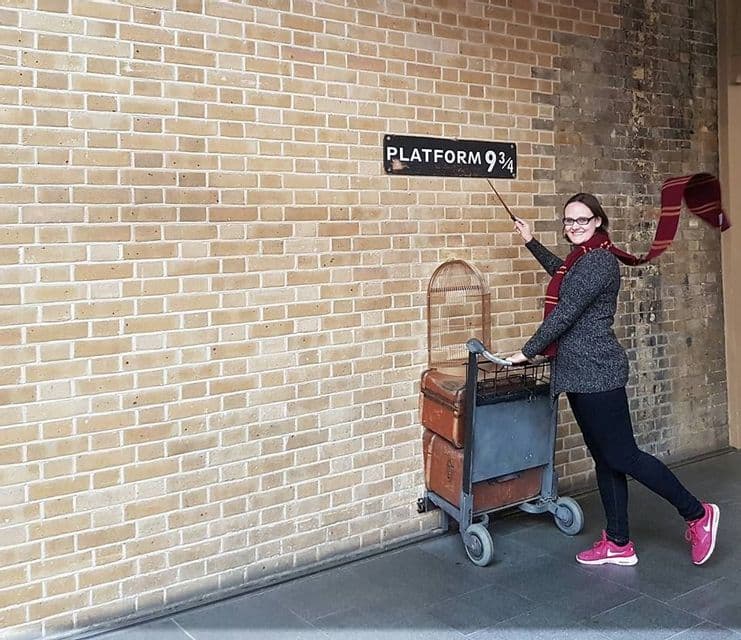 Eine Frau mit gestreiftem Schal posiert mit Zauberstab und Gepäckwagen am Schild 'Gleis 9¾' an einer Backsteinmauer.