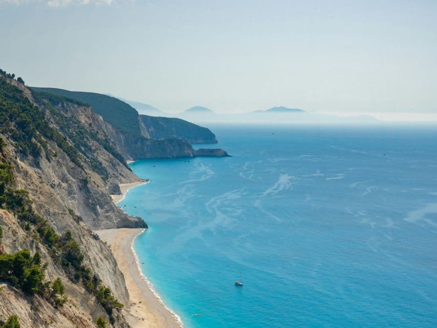 Veduta dall'alto di una costa frastagliata, con una spiaggia sabbiosa incastonata tra le scogliere e una barca a vela sul mare turchese.