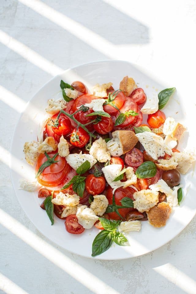 Una vista dall'alto di un'insalata fresca con pomodori, mozzarella, crostini e foglie di basilico su un piatto bianco sotto la luce solare a strisce.