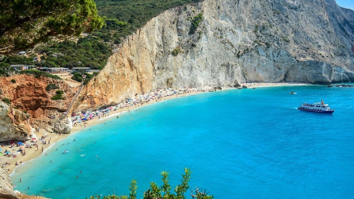 Una vista dall'alto di una spiaggia affollata con persone che nuotano in acque di un turchese brillante, alla base di una grande scogliera calcarea bianca.