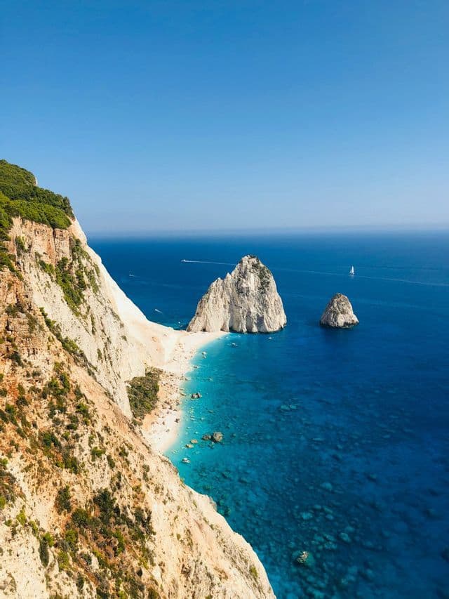 Una vista dall'alto di una scogliera ripida e rocciosa che si affaccia su una spiaggia isolata di sabbia bianca con due grandi faraglioni nell'acqua turchese.