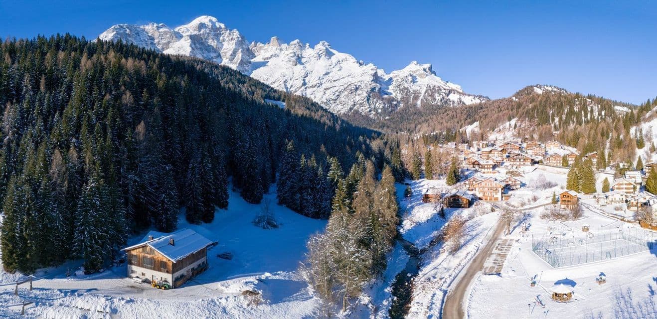 Una vista aerea di un villaggio innevato con chalet in legno annidati in una pineta ai piedi di una catena montuosa frastagliata.
