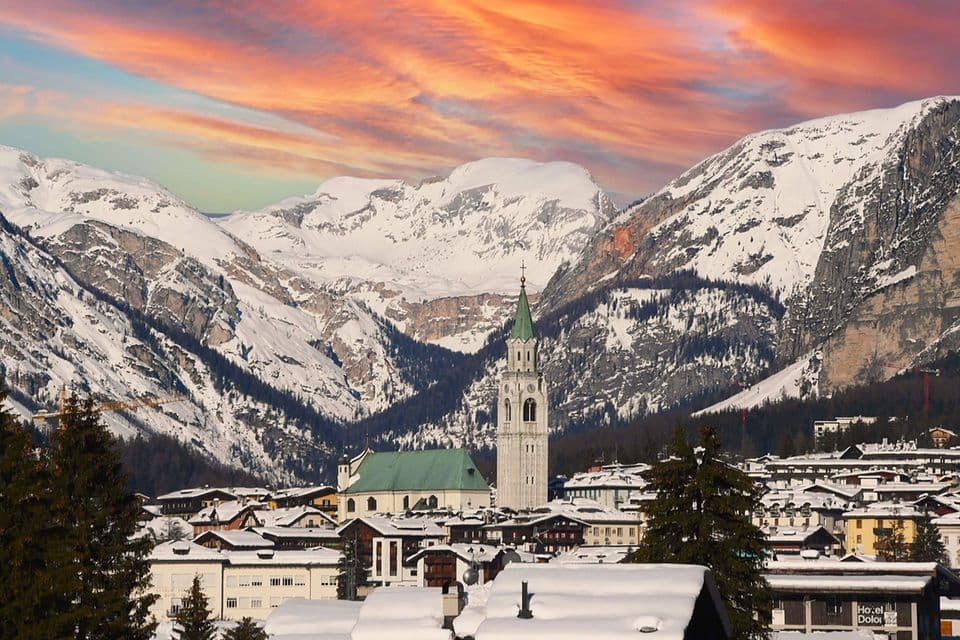 Un villaggio innevato con una chiesa imponente è adagiato tra grandi montagne innevate sotto un cielo colorato al tramonto.