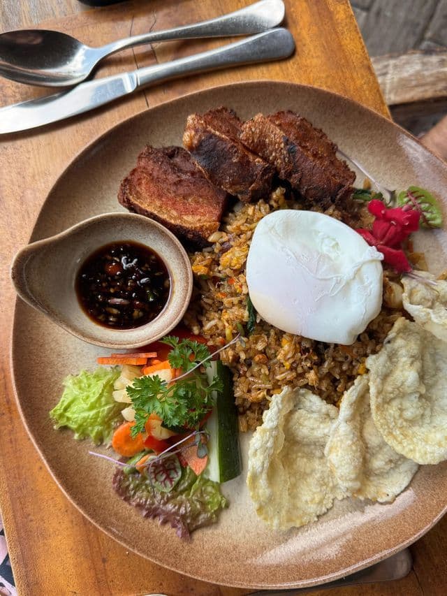 Un piatto di riso fritto con un uovo in camicia, servito con pancetta di maiale croccante, cracker, verdure fresche e una salsa da intingere.