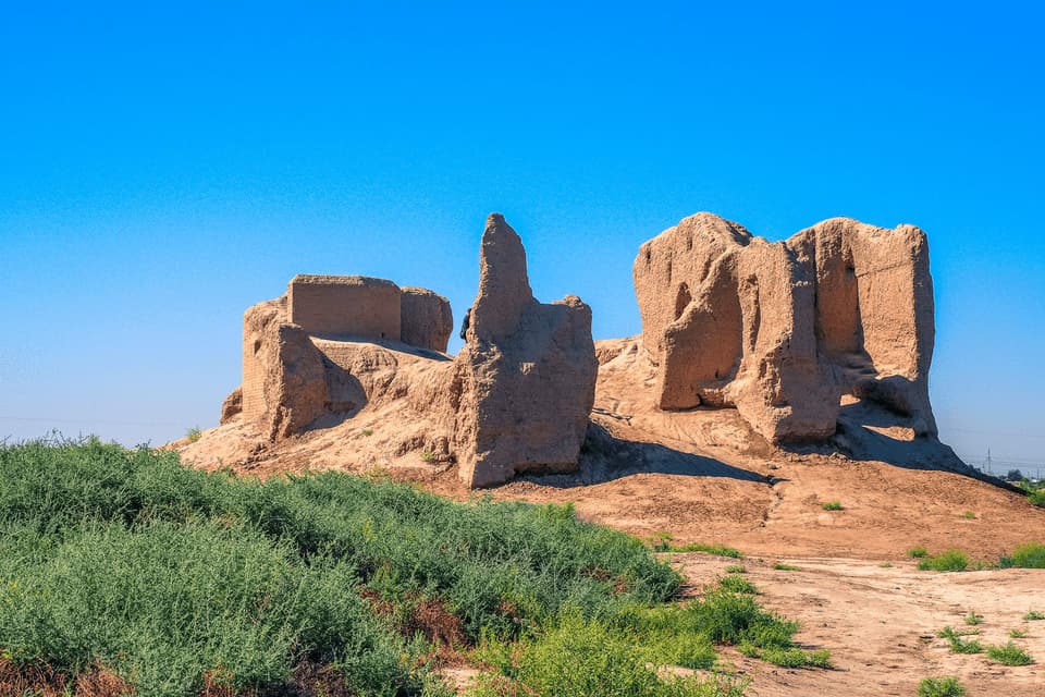 Antiche rovine di mattoni di fango si ergono su una collina sabbiosa sotto un cielo azzurro limpido, con cespugli verdi in primo piano.