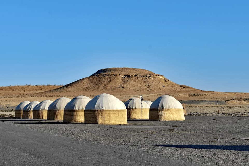 Una fila di yurte con basi intrecciate si trova in un paesaggio desertico con una grande collina sullo sfondo sotto un cielo azzurro chiaro.