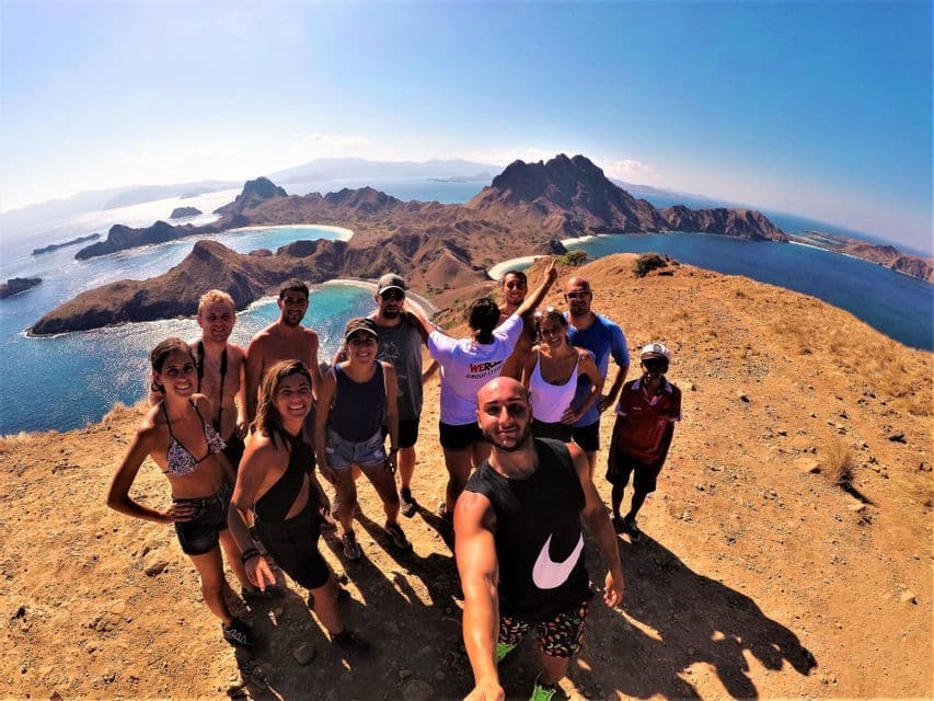 Un voyage de groupe WeRoad prend un selfie grand-angle sur une colline ensoleillée surplombant une côte spectaculaire avec des îles et une eau bleue.