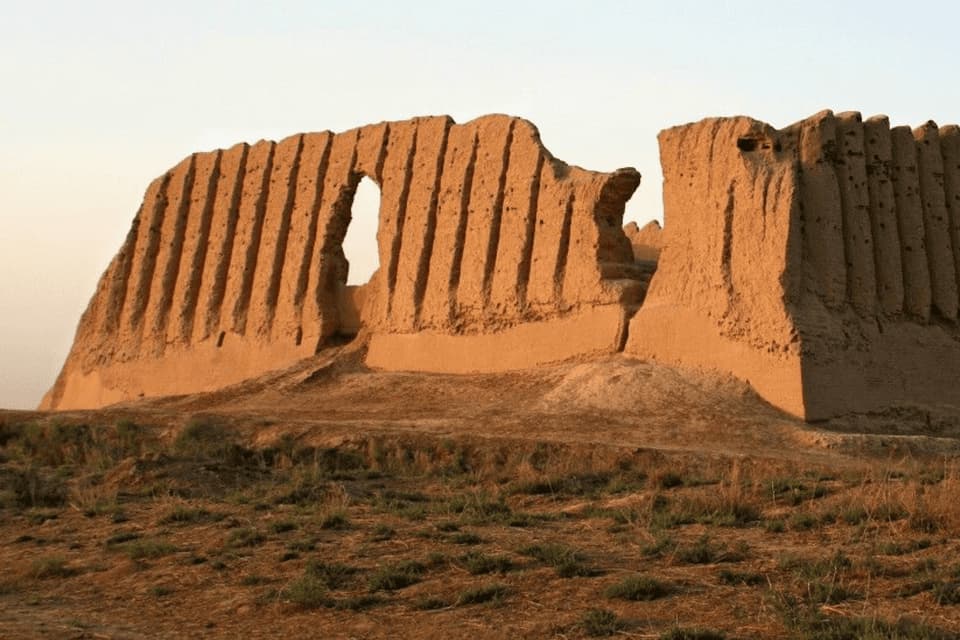 Le antiche rovine fatiscenti di un muro di fortezza in mattoni di fango si ergono su una collina arida ed erbosa sotto un cielo pallido.
