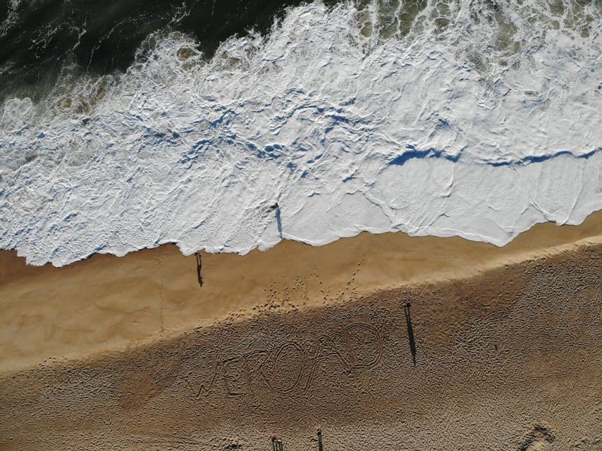 An aerial view of a sandy beach with 'WEROAD' written in the sand, while people stand near the shoreline as a wave crashes.
