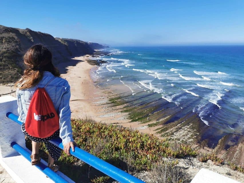 Une femme avec un sac à dos rouge WeRoad s'appuie sur une balustrade bleue, regardant une côte sablonneuse avec des vagues et des falaises.