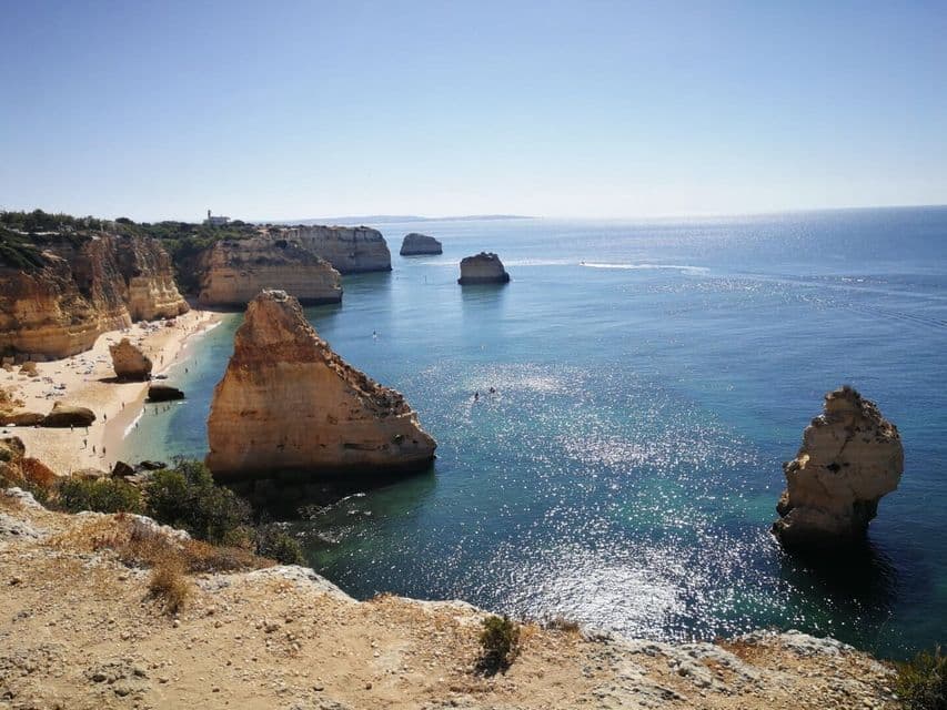 Un littoral de falaises abruptes, une plage de sable animée, et de grandes formations rocheuses émergeant de la mer d'un bleu étincelant.