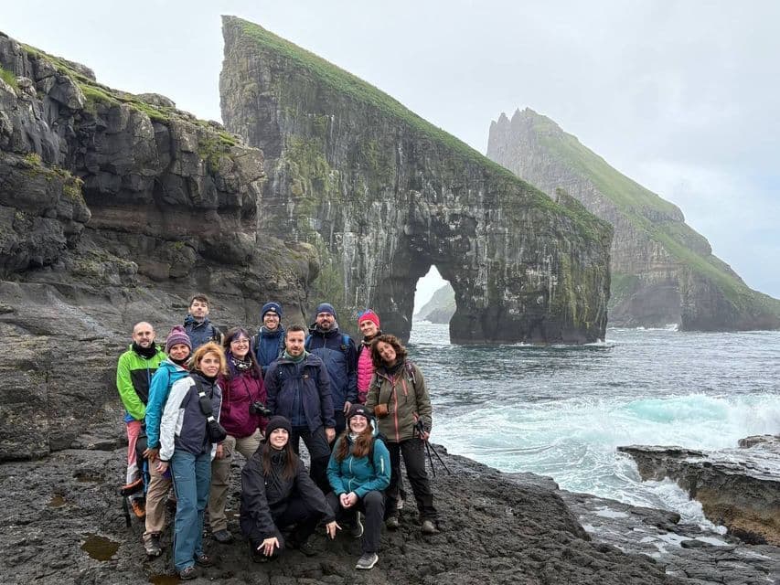 Un gruppo WeRoad in posa su una costa rocciosa con una grande scogliera e un arco naturale sullo sfondo sotto un cielo coperto.