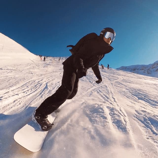 Ein Snowboarder mit Helm und schwarzer Ausrüstung zieht Spuren in einen sonnenbeschienenen, schneebedeckten Hang unter klarem blauen Himmel.