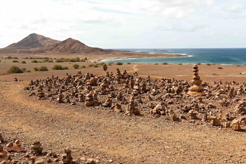 De nombreux petits cairns de pierres équilibrées parsèment un paysage côtier aride, avec des montagnes et l'océan en arrière-plan.