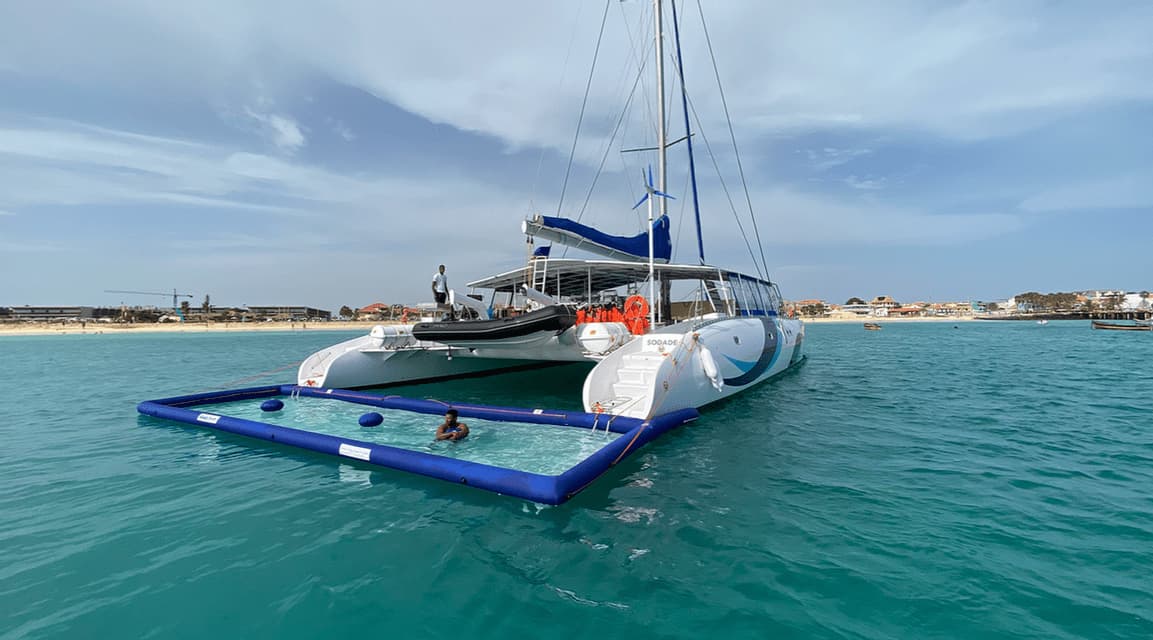 Un catamaran ancré dans des eaux turquoise avec une personne nageant dans une piscine flottante attenante près d'une plage de sable.