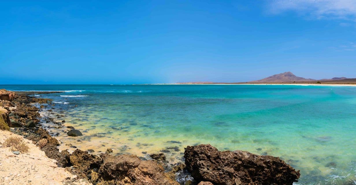 Une vue panoramique d'une côte rocheuse avec une eau turquoise claire, menant à une plage et une montagne lointaines sous un ciel bleu éclatant.