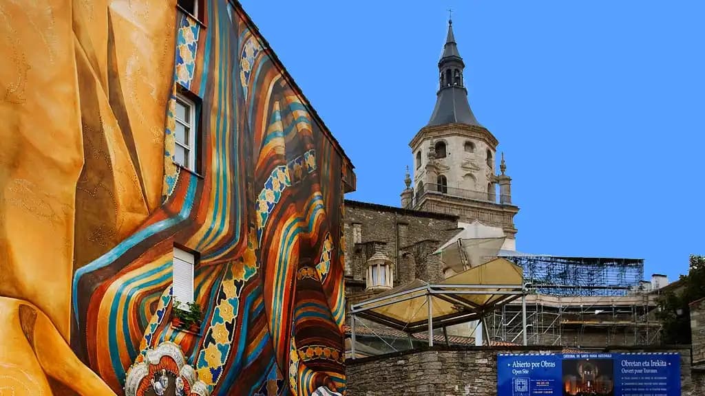 Un murale colorato e moderno su una facciata di un edificio si erge accanto a un'antica torre campanaria in pietra sotto un cielo blu limpido.