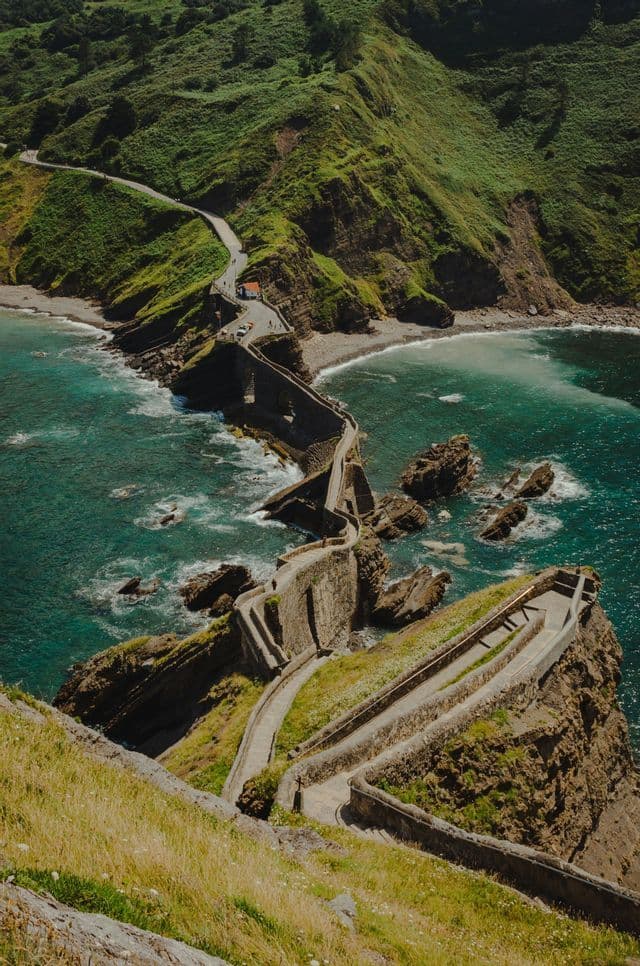 Un sentiero lastricato tortuoso con scalini conduce da una costa verde e collinare a un piccolo isolotto roccioso circondato da acque turchesi.