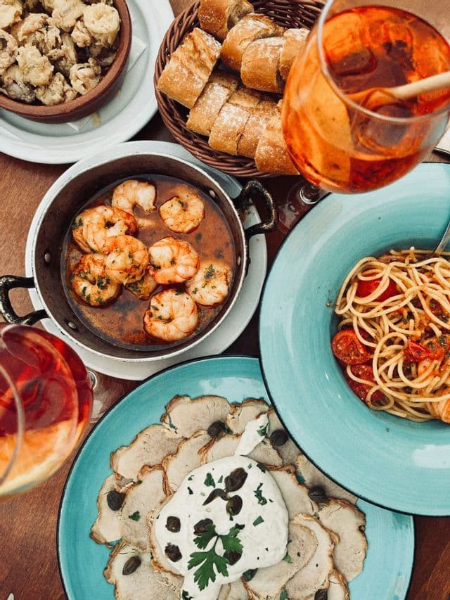 Una vista dall'alto di un tavolo imbandito con vari piatti come gamberi in padella, spaghetti, carne affettata con salsa e cocktail.