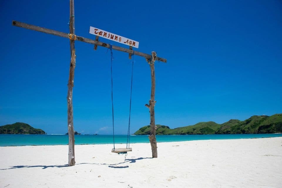 Eine Holzschaukel an einem weißen Sandstrand mit Blick auf ein türkisfarbenes Meer und grüne Hügel im Hintergrund.