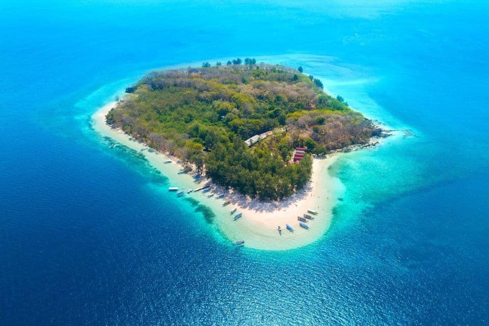 Eine Luftaufnahme einer kleinen tropischen Insel mit einem weißen Sandstrand, umgeben von klarem türkisblauem und tiefblauem Ozean.