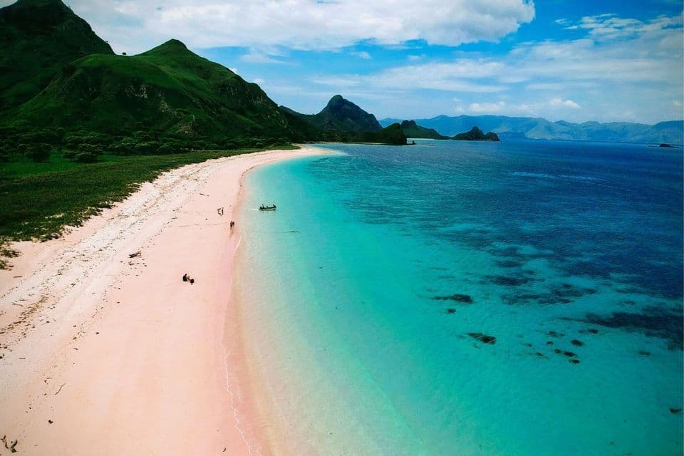 Eine Luftaufnahme eines rosa Sandstrandes neben türkisfarbenem Wasser mit üppig grünen Bergen im Hintergrund.