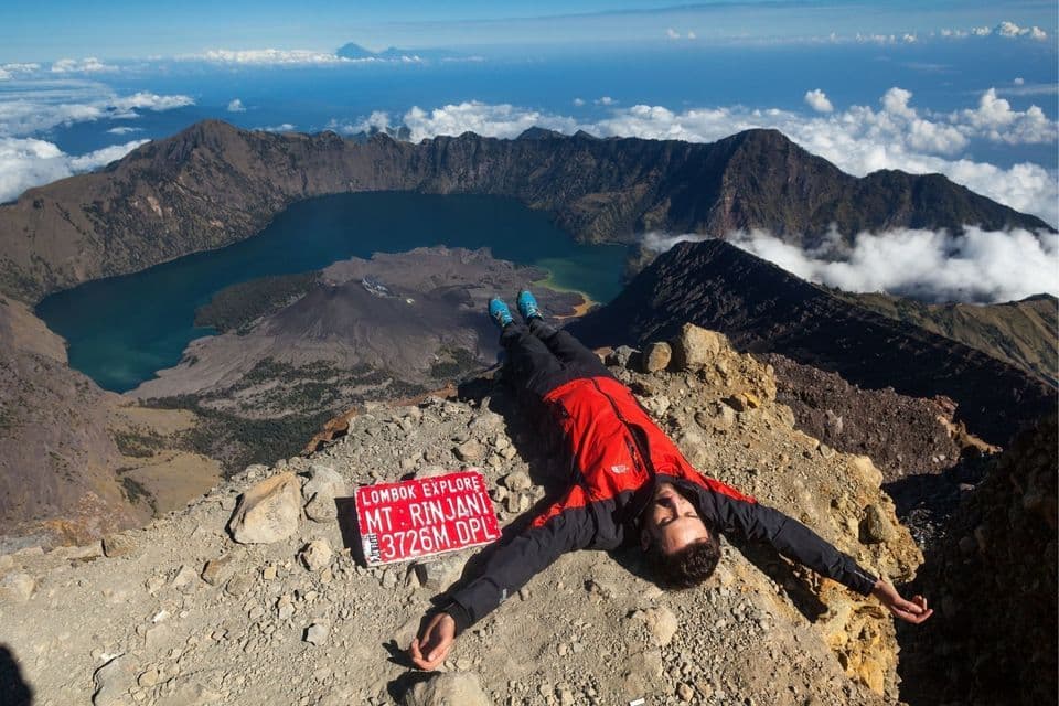 Ein Wanderer in roter Jacke liegt auf dem Rücken auf dem Gipfel des Mount Rinjani, mit Vulkankrater und See im Hintergrund.