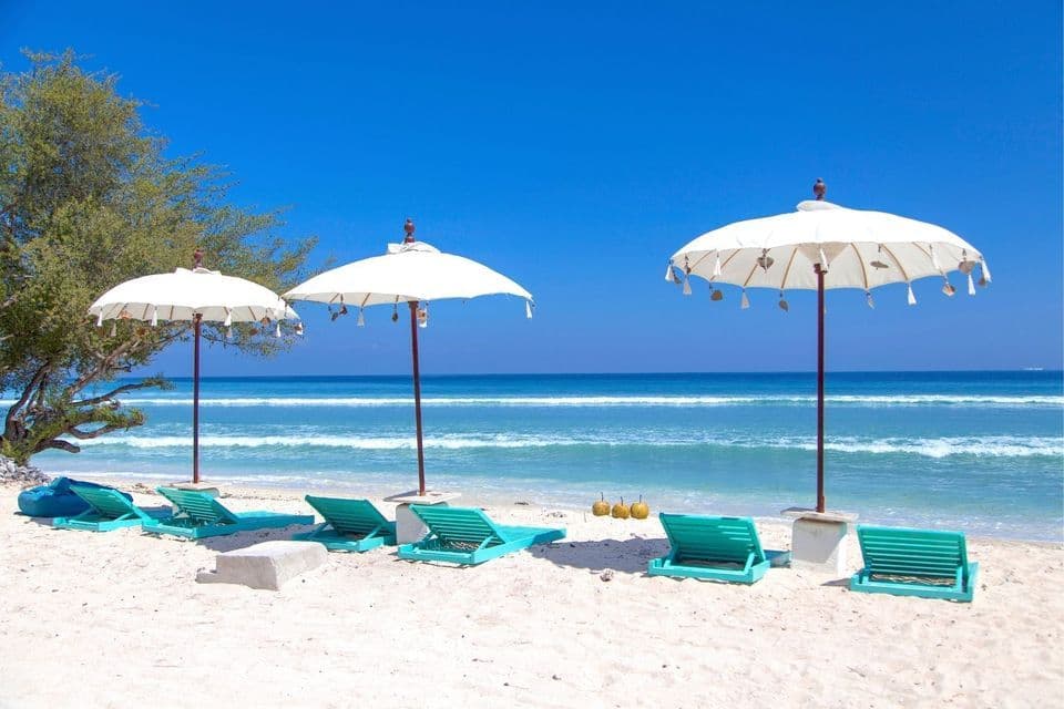 Eine Reihe türkisfarbener Liegestühle und weißer Sonnenschirme an einem Sandstrand mit Blick auf einen ruhigen, blauen Ozean unter klarem Himmel.