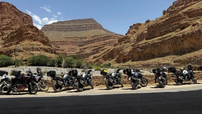 Una fila di motociclette da turismo parcheggiate a bordo strada, ai piedi di un grande canyon roccioso a strati.