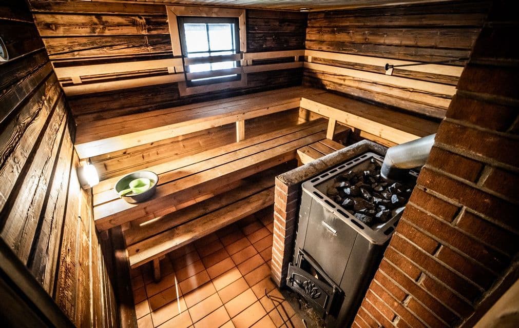 Das Innere einer traditionellen Holzsauna mit gestuften Bänken, einem gefliesten Boden und einem Ofen mit heißen Steinen.