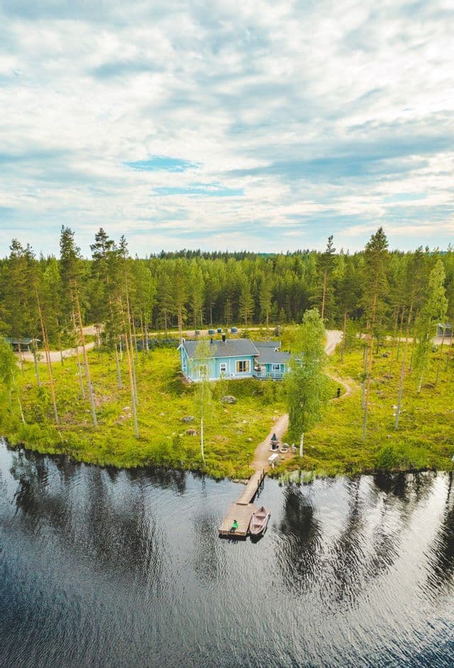 Eine Luftaufnahme eines blauen Holzhauses an einem grasbewachsenen Seeufer, mit einem Steg, zwei kleinen Booten und einem umliegenden Kiefernwald.