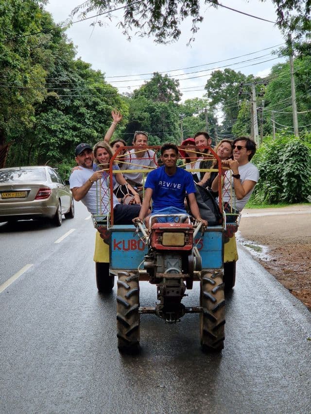 Un gruppo WeRoad sorride mentre viaggia sul retro di un piccolo trattore blu su una strada bagnata fiancheggiata da alberi.