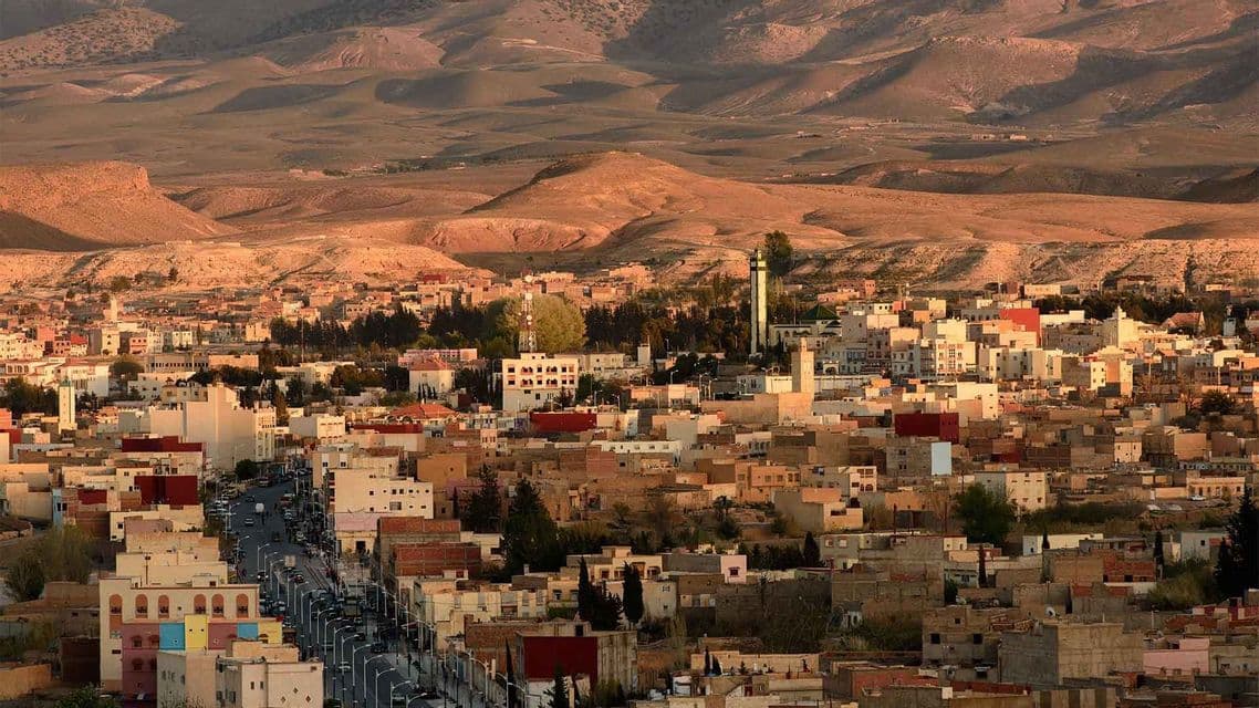 Una vista panoramica di una città densamente popolata con edifici color terra, ai piedi di grandi colline desertiche ondulate, al tramonto.