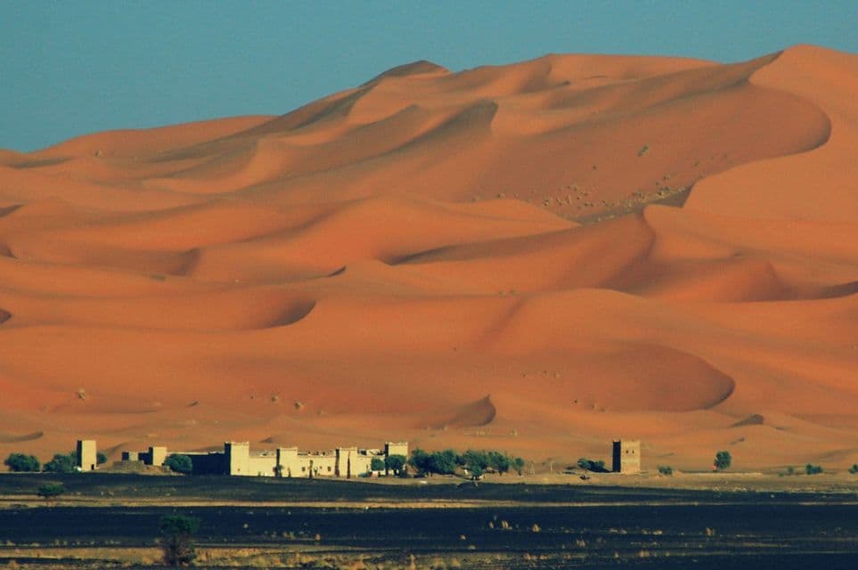 Un piccolo insediamento di edifici in adobe e alberi si trova alla base di imponenti dune di sabbia arancione sotto un cielo azzurro e limpido.