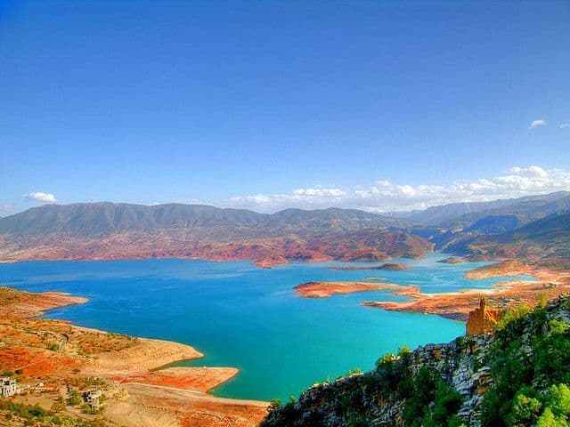 Una vista panoramica su un lago turchese brillante, circondato da colline bruno-rossastre e montagne lontane, sotto un cielo azzurro e limpido.