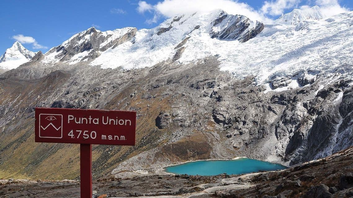 Un cartello rosso per Punta Union si erge di fronte a un lago alpino turchese e montagne innevate sotto un cielo azzurro.