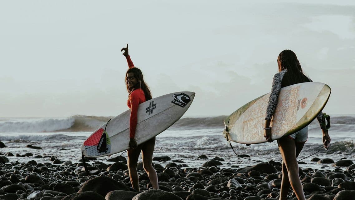 Due donne di un viaggio di gruppo WeRoad trasportano tavole da surf su una spiaggia rocciosa, e una sorride alla fotocamera in riva all'oceano.