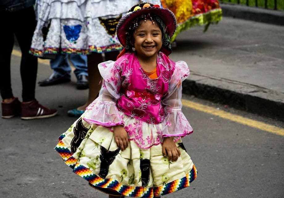 Una ragazza sorride, indossando un abito tradizionale colorato e un cappello, su una strada lastricata durante un festival.