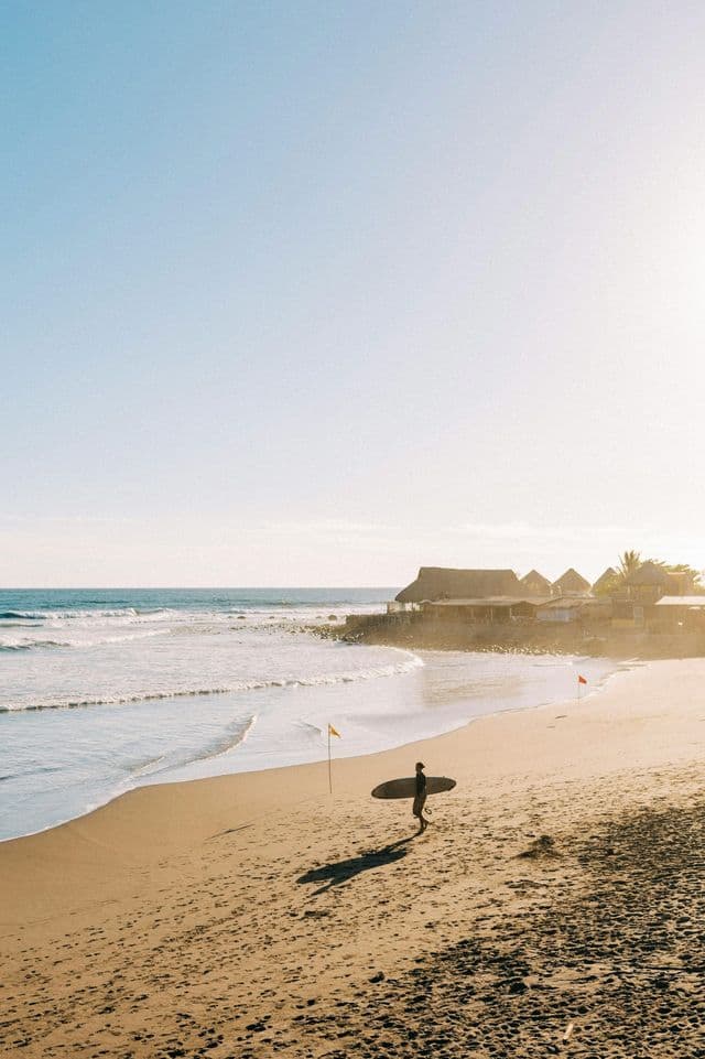 Un surfista in controluce porta la sua tavola camminando su una spiaggia sabbiosa vicino all'oceano all'alba.