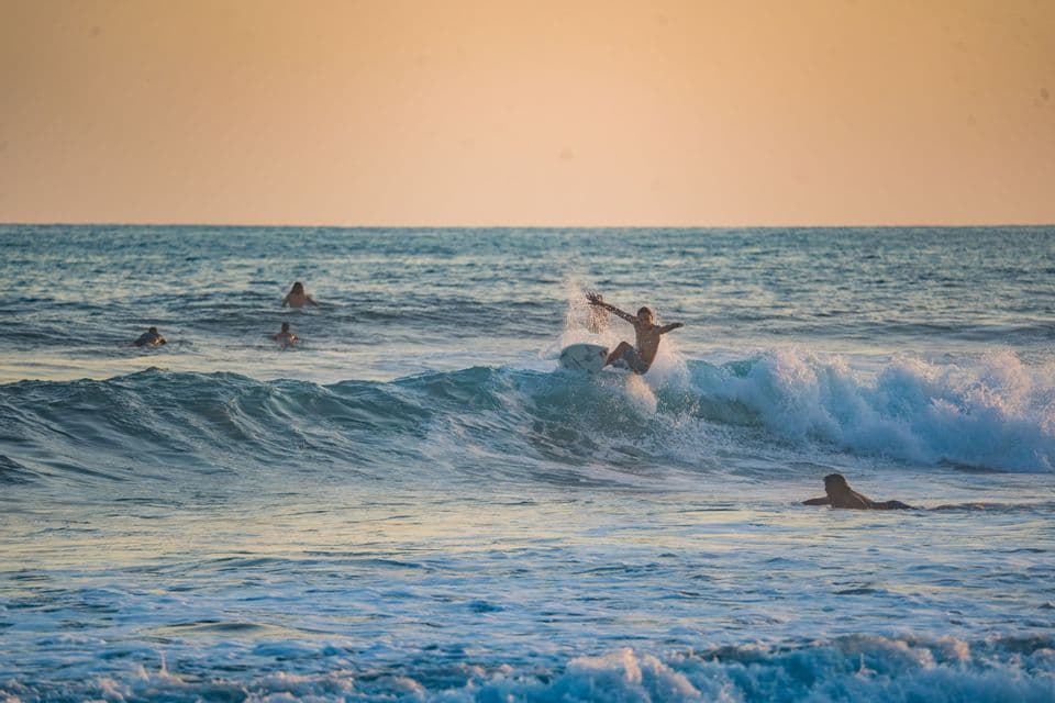 Un surfista su un'onda imponente, schizzando acqua mentre altre persone nuotano nell'oceano al tramonto.